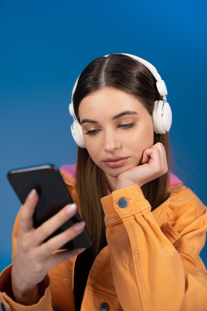 Teen girl in headphones using blurred smartphone isolated on blue  - Fotoğraf, Görsel