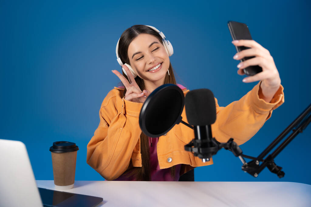 Teenager in headphones taking selfie on smartphone near studio microphone and laptop isolated on blue  - Fotografie, Obrázek