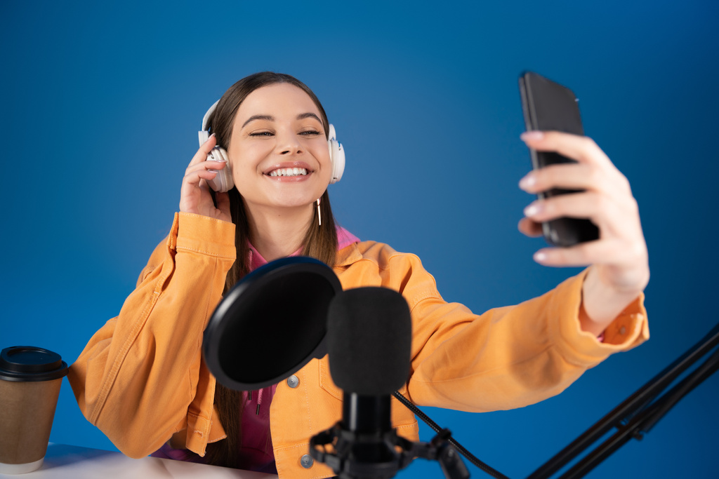 Smiling teenager in headphones taking selfie on cellphone near studio microphone isolated on blue  - Photo, image