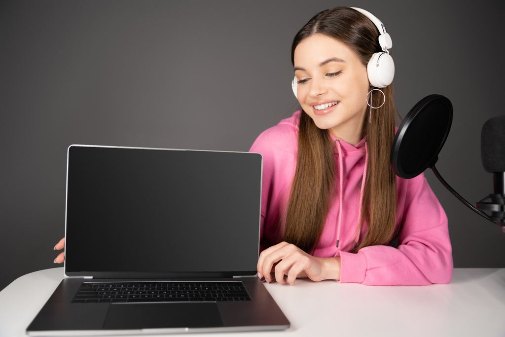 Hymyilevä teini podcaster tarkastellaan kannettava tietokone tyhjä näyttö lähellä mikrofonia eristetty harmaa  - Valokuva, kuva