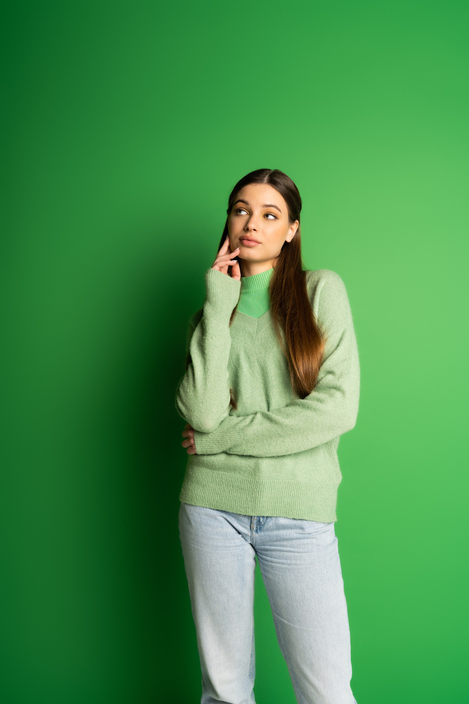 Pensive teen girl in jumper and jeans looking away on green background  - Photo, Image