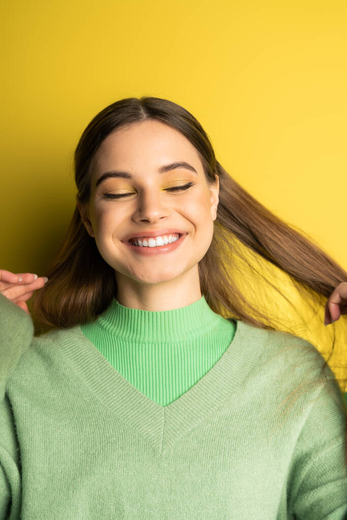 Cheerful teenager in jumper touching hair on yellow background  - Foto, Imagen