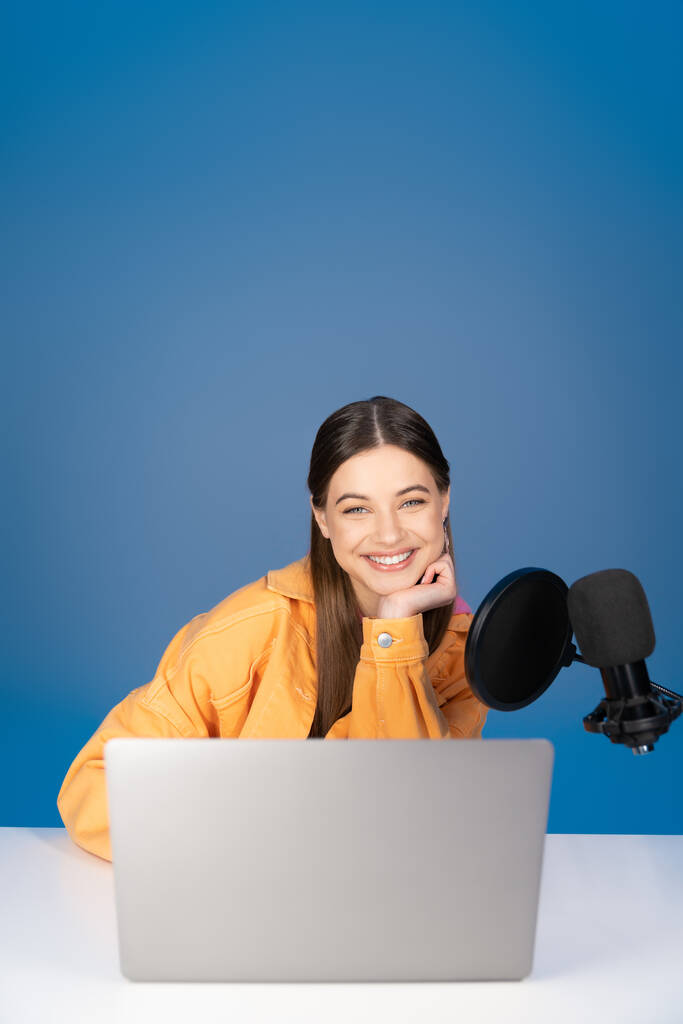Χαρούμενος έφηβος με πορτοκαλί μπουφάν χαμογελώντας στην κάμερα κοντά στο φορητό υπολογιστή και το μικρόφωνο του στούντιο που απομονώνονται σε μπλε  - Φωτογραφία, εικόνα