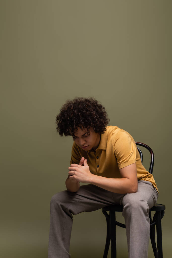 curly african american man in yellow polo shirt and trousers sitting on chair with bowed head on grey green background - Photo, Image