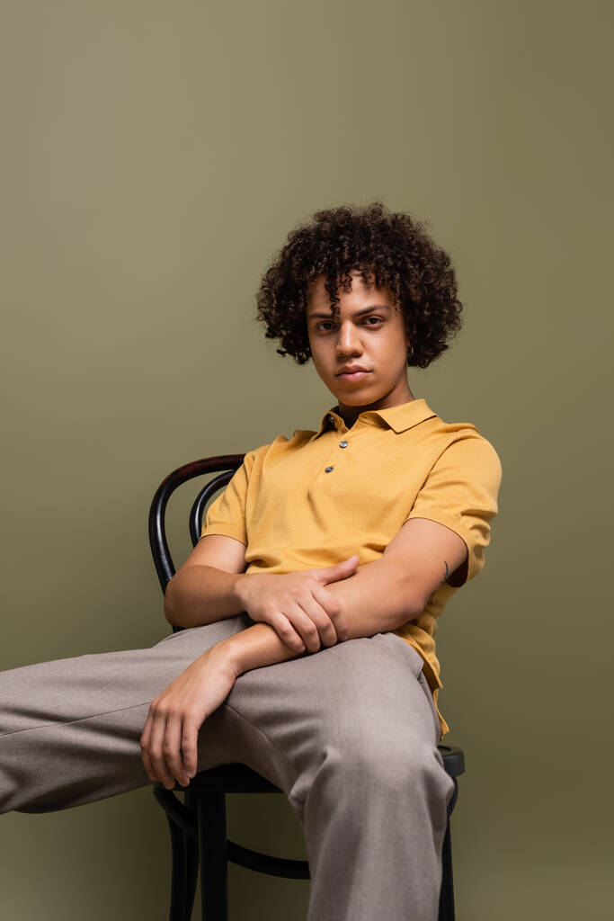 trendi afro-amerikai férfi sárga pólóban ül a széken, és nézi a kamera szürke háttér - Fotó, kép