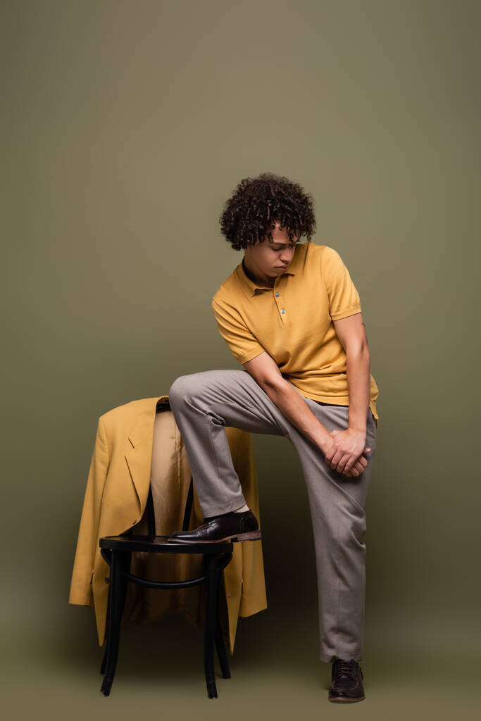 trendy uomo afroamericano passo sulla sedia con giacca gialla mentre posa su sfondo grigio verde - Foto, immagini