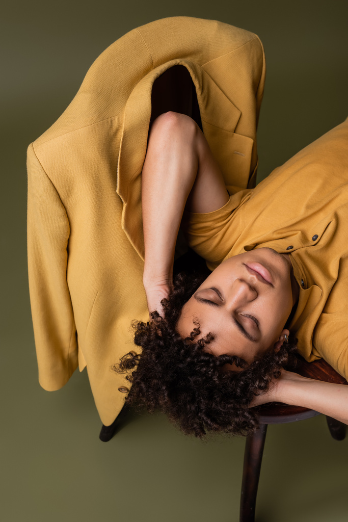 high angle view of african american man lying with closed eyes and hands behind head near yellow blazer on chair on grey green background - 写真・画像