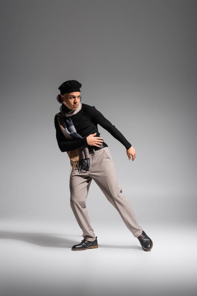 full length of african american man in stylish beret and black sweater with checkered scarf looking away while posing on grey background - Photo, Image