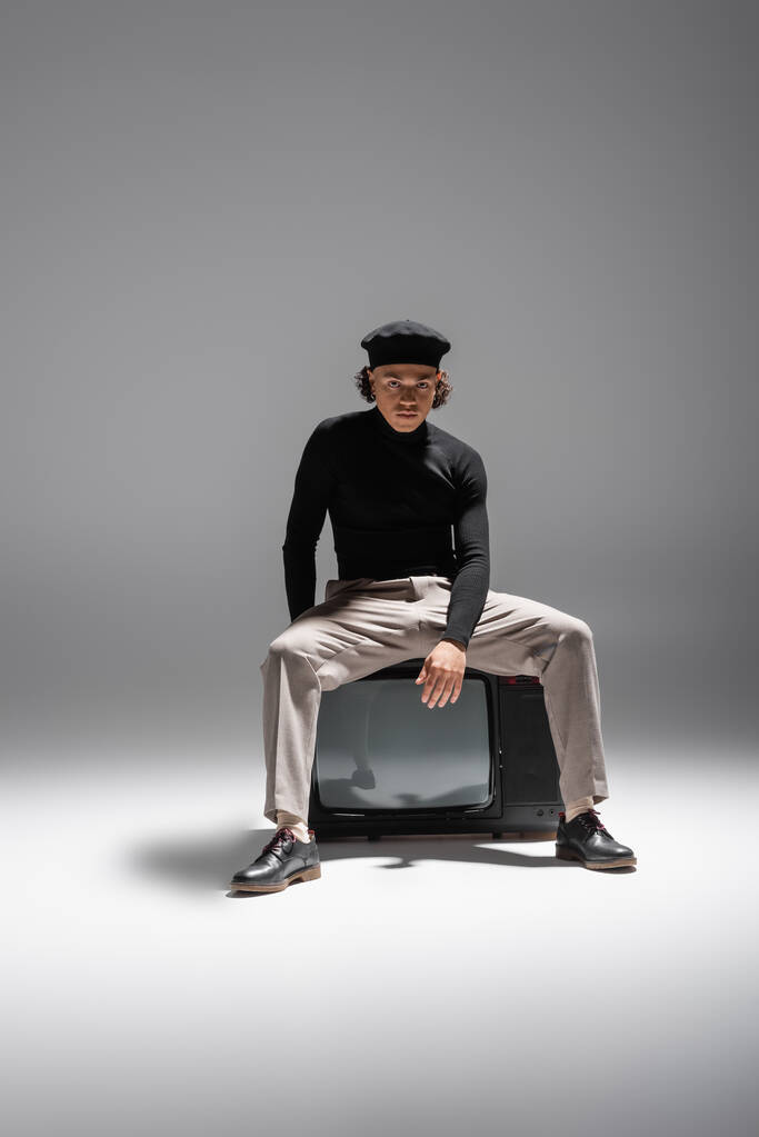 full length of trendy african american man in black turtleneck and pants with leather shoes sitting on vintage tv set on grey background - 写真・画像