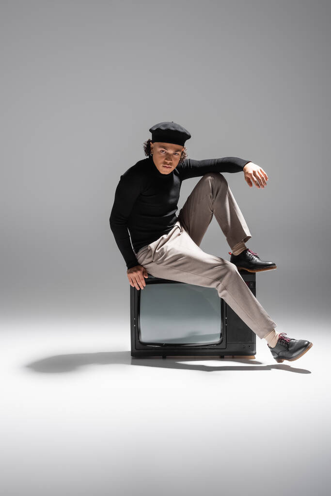 full length of african american man in stylish beret and black pullover sitting on retro tv and looking at camera on grey background - Φωτογραφία, εικόνα