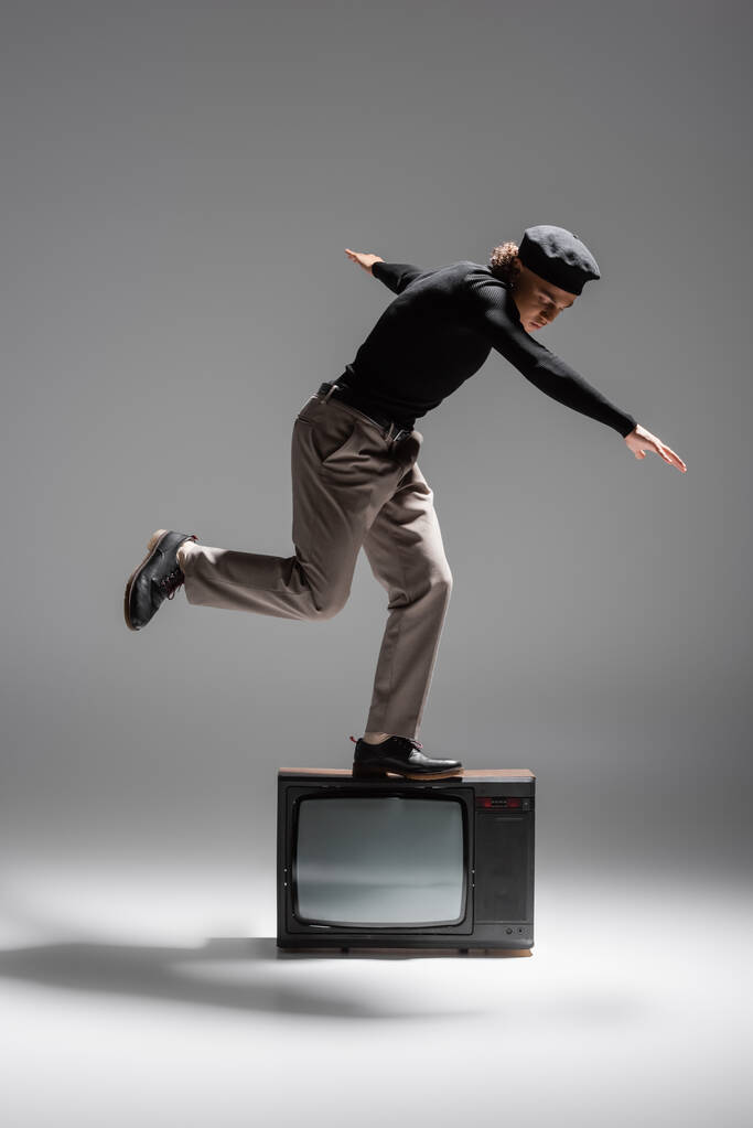 full length of trendy african american man in black sweater and trousers balancing on vintage tv set on grey background - Foto, Bild