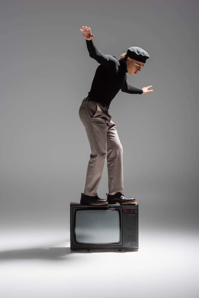 volle Länge der afrikanisch-amerikanischen Mann in trendiger Freizeitkleidung Balancieren auf Vintage-TV-Set auf grauem Hintergrund - Foto, Bild