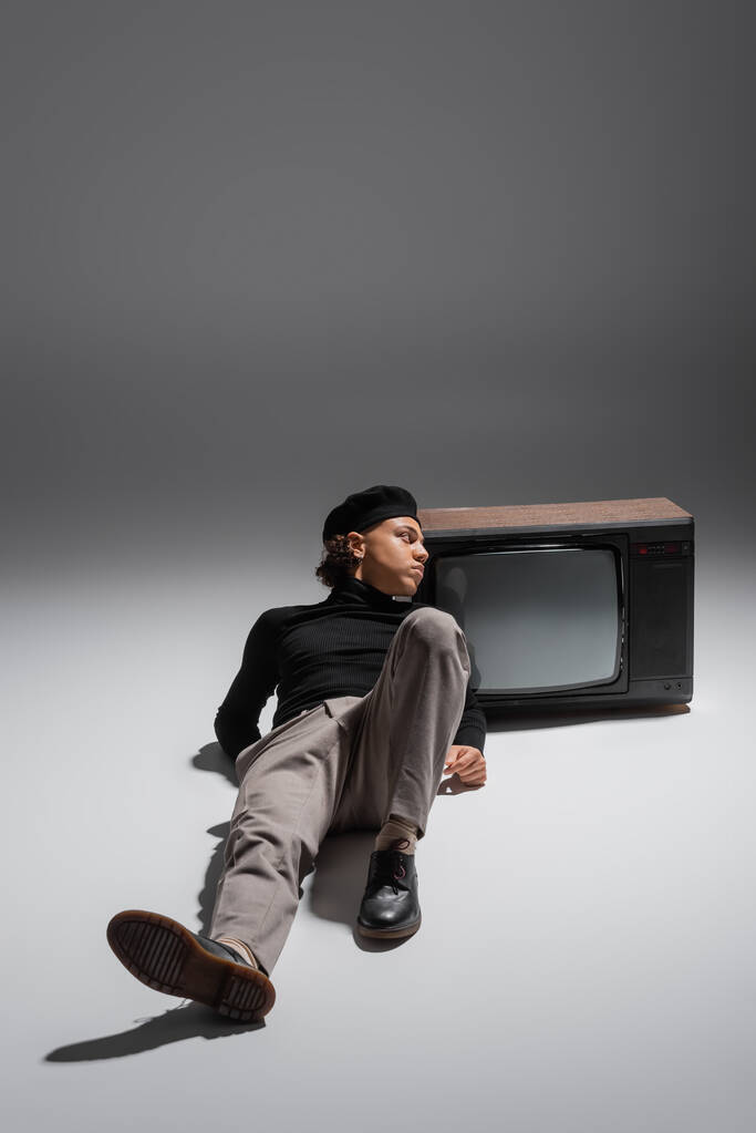 comprimento total do homem americano africano na moda em gola alta preta e boina deitada perto do aparelho de TV vintage e olhando para o cinza  - Foto, Imagem