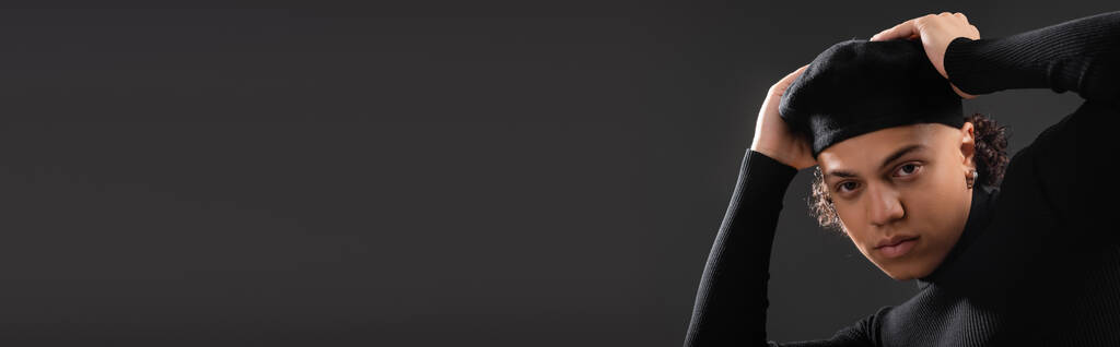 молодий афроамериканський чоловік в чорному светрі регулює стильний берет, дивлячись на камеру ізольовано на темно-сірому, банер
 - Фото, зображення