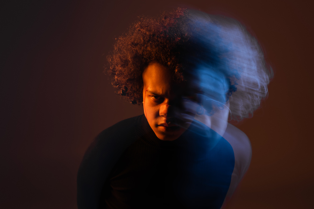 larga exposición del hombre afroamericano herido con trastorno bipolar mirando a la cámara sobre fondo oscuro con luz naranja y azul - Foto, Imagen