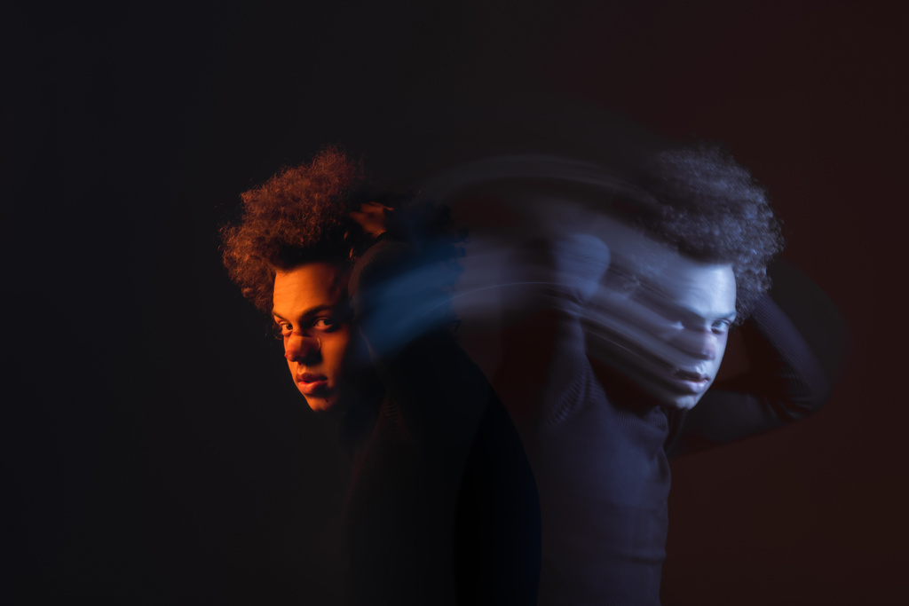 dvojitá expozice zraněného afrického Američana s bipolární poruchou při pohledu na kameru na tmavém pozadí s oranžovým a modrým světlem - Fotografie, Obrázek