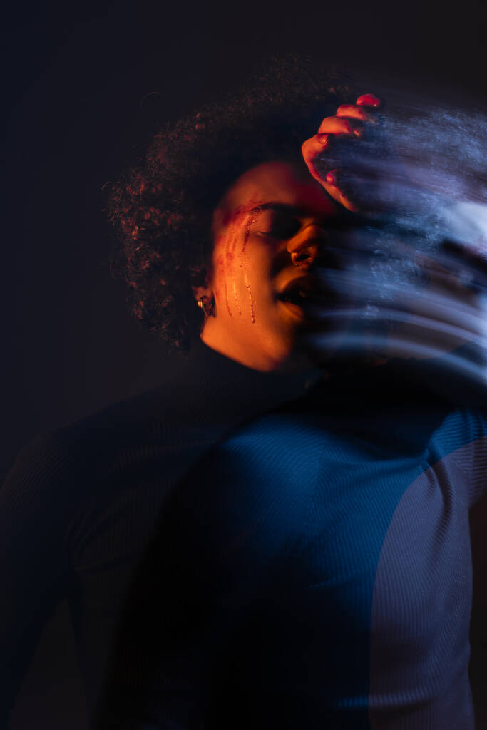διπλή έκθεση του τραυματισμένου Αφροαμερικανού άνδρα στέκεται με το χέρι κοντά στο πρόσωπο αιμορραγία σε σκούρο φόντο με κόκκινο και μπλε φως - Φωτογραφία, εικόνα