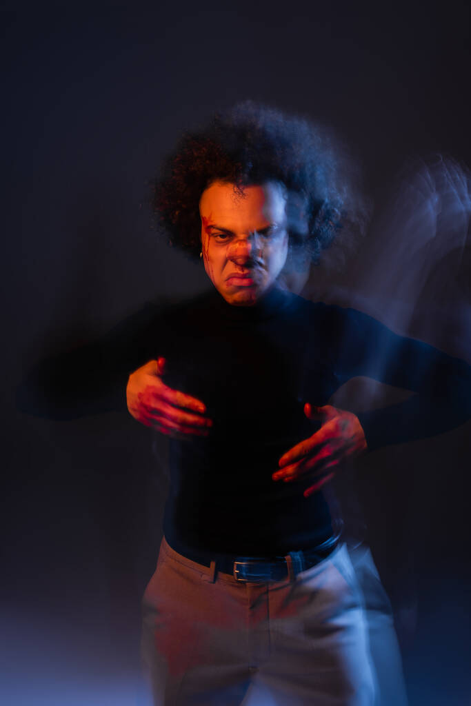 bewegingsvervaging van agressieve Afro-Amerikaanse man met bipolaire stoornis en bloederig gezicht kijkend naar camera op donker met oranje en blauw licht - Foto, afbeelding