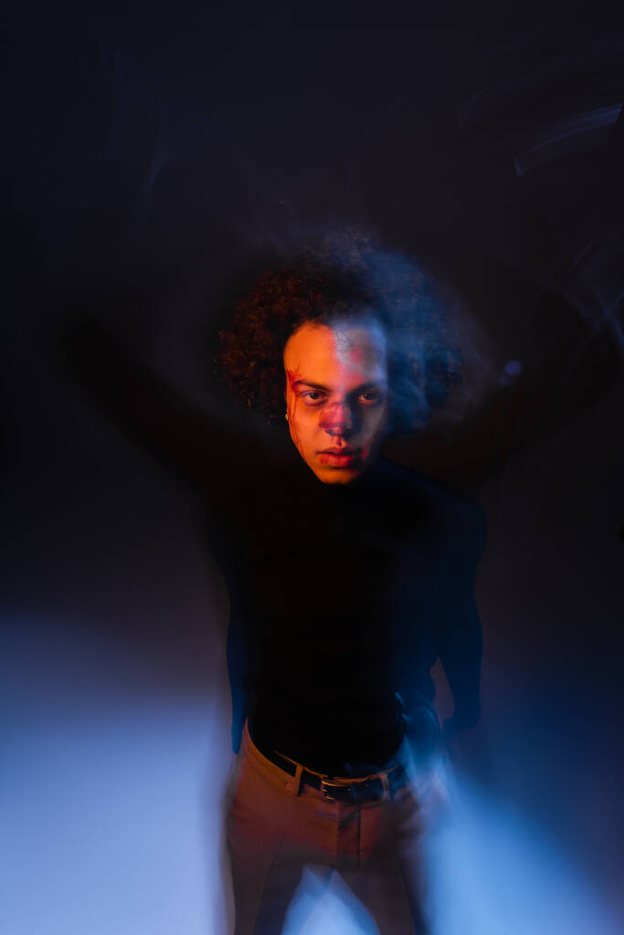 homem americano africano ferido com transtorno bipolar e sangramento rosto olhando para a câmera no fundo escuro com luz laranja e azul - Foto, Imagem