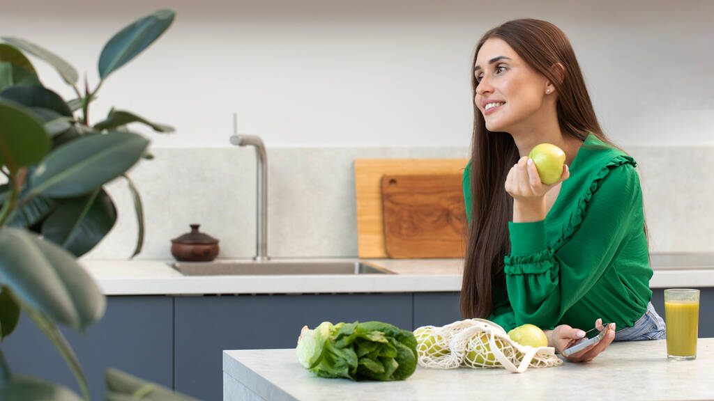 Onnellinen nuori nainen pitää vihreää omenaa kädessään keittiössä. Käsite terveellistä ruokavaliota, kasvissyöntiä. - Valokuva, kuva