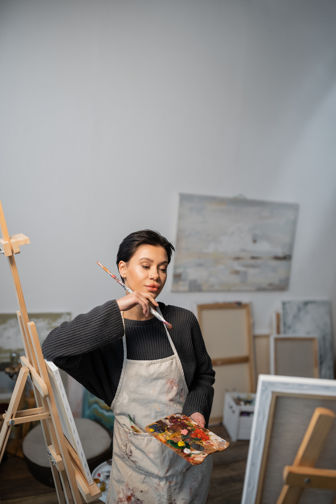 Focused artist holding palette and looking at painting in workshop  - Zdjęcie, obraz