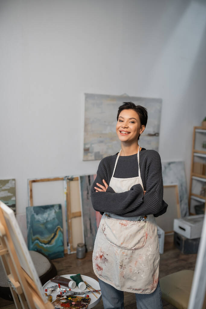 Cheerful artist in apron looking at camera near paints and easel in studio  - Foto, Bild
