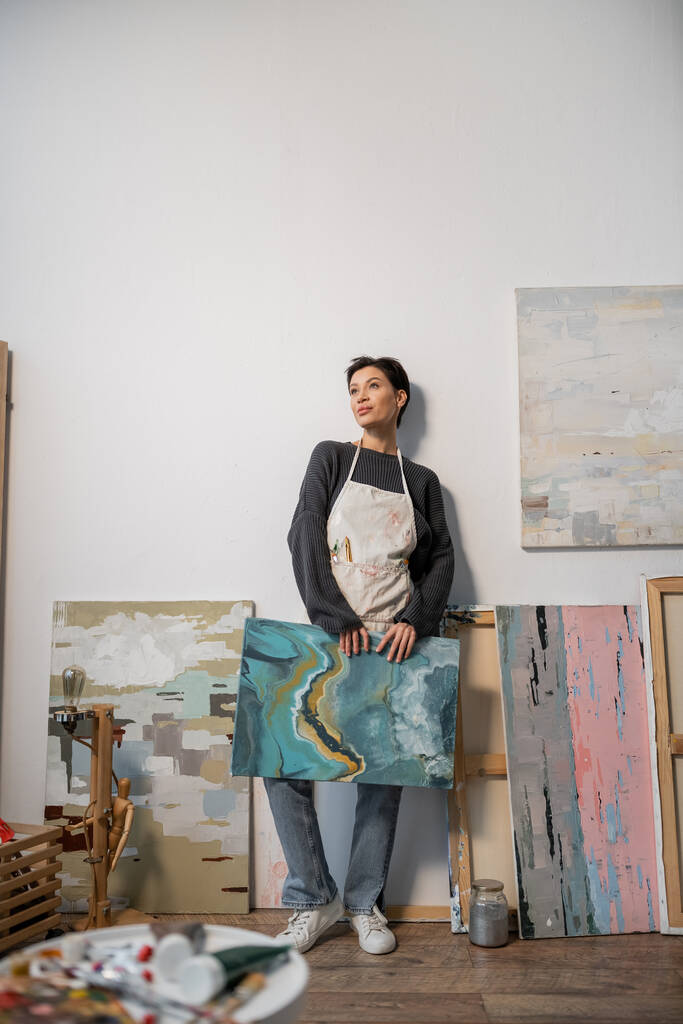 Брюнетка в фартуке держит рисунок рядом с размытыми красками в мастерской  - Фото, изображение