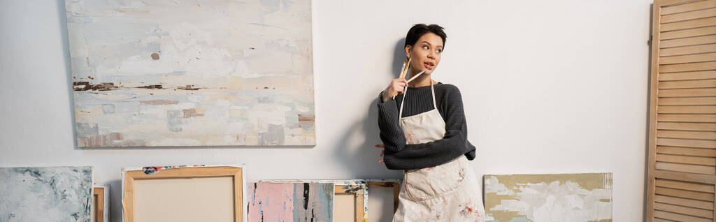 Мечтательный молодой художник держит кисти возле рисунков в мастерской, баннер  - Фото, изображение