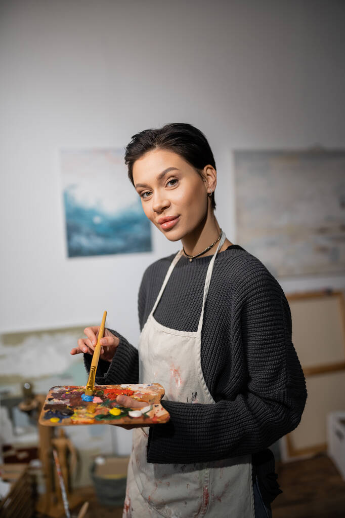 Portrait of short haired artist in apron holding paintbrush and palette in workshop  - Fotó, kép