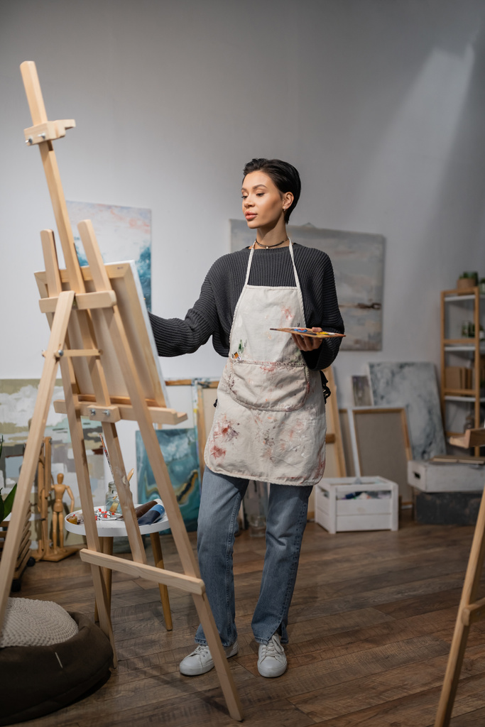 Artist in apron holding palette and looking at canvas on easel in studio  - Foto, Imagen