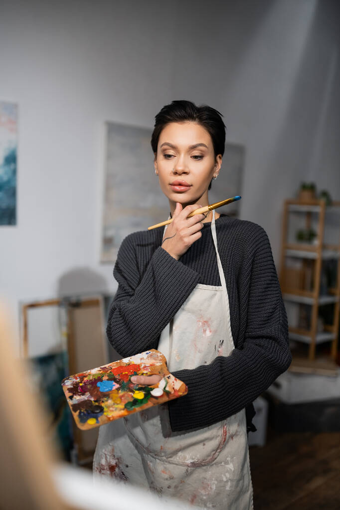 Концентрированный художник держит кисть и палитру в мастерской  - Фото, изображение