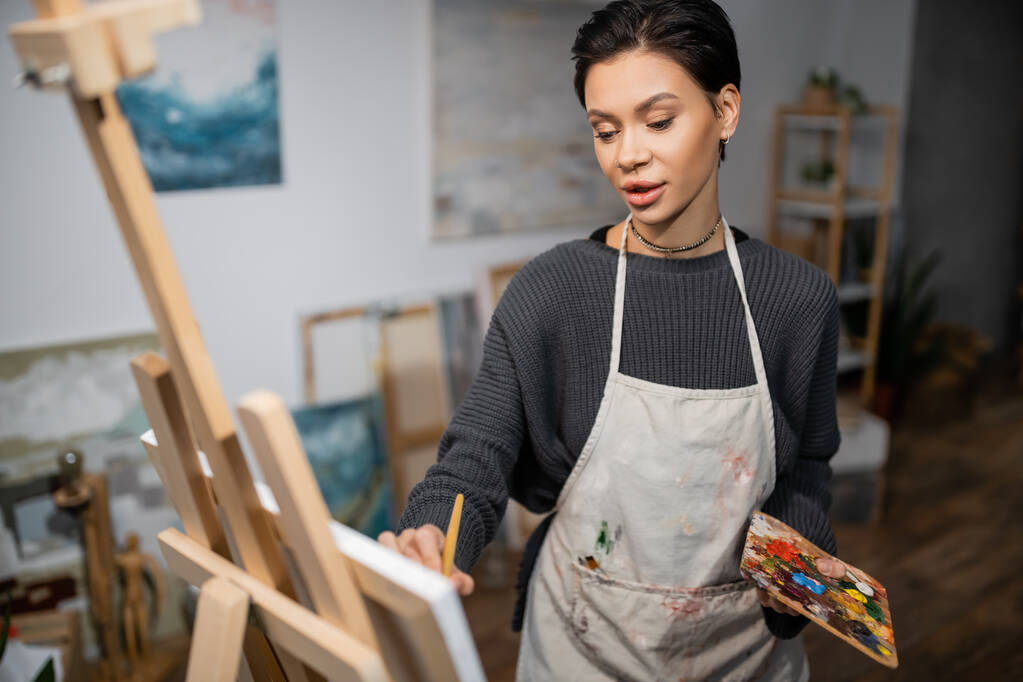 Short haired artist in apron holding palette while painting on blurred canvas  - Foto, afbeelding