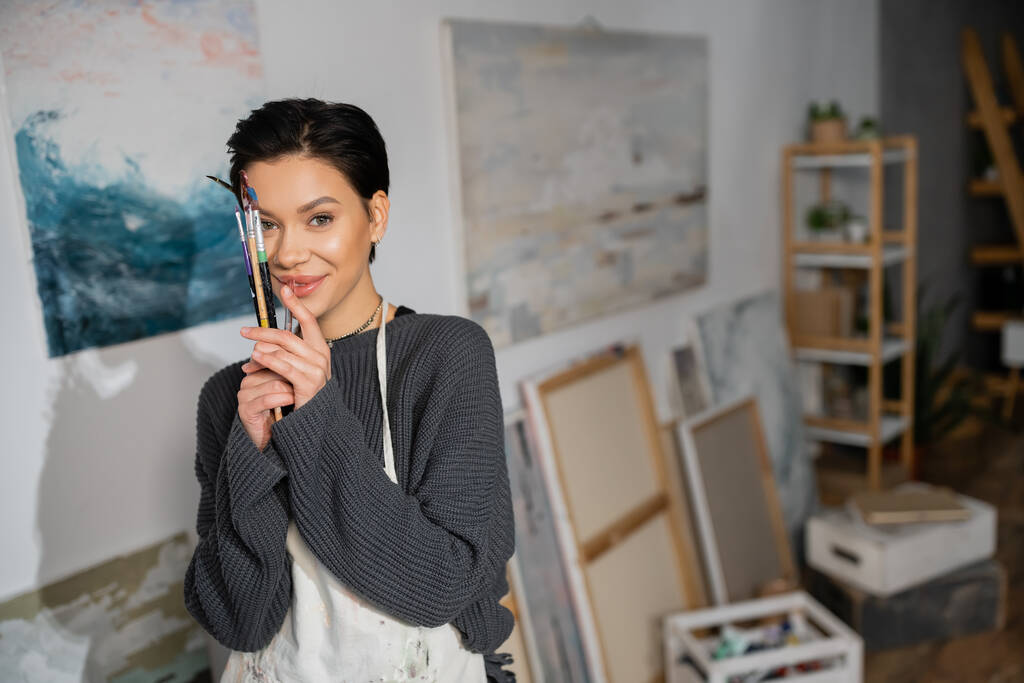 Smiling artist in apron holding paintbrushes near face in studio  - Fotografie, Obrázek