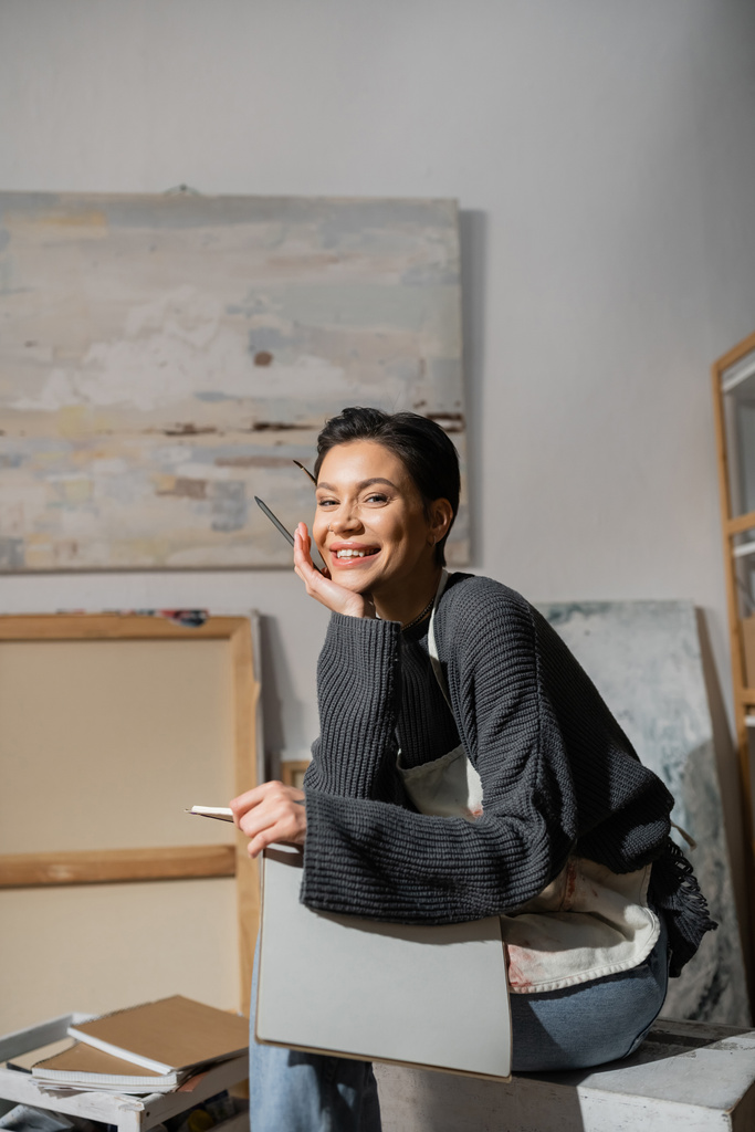 Χαρούμενος καλλιτέχνης με ποδιά κρατώντας μπλοκ ζωγραφικής και κοιτάζοντας την κάμερα στο εργαστήριο  - Φωτογραφία, εικόνα