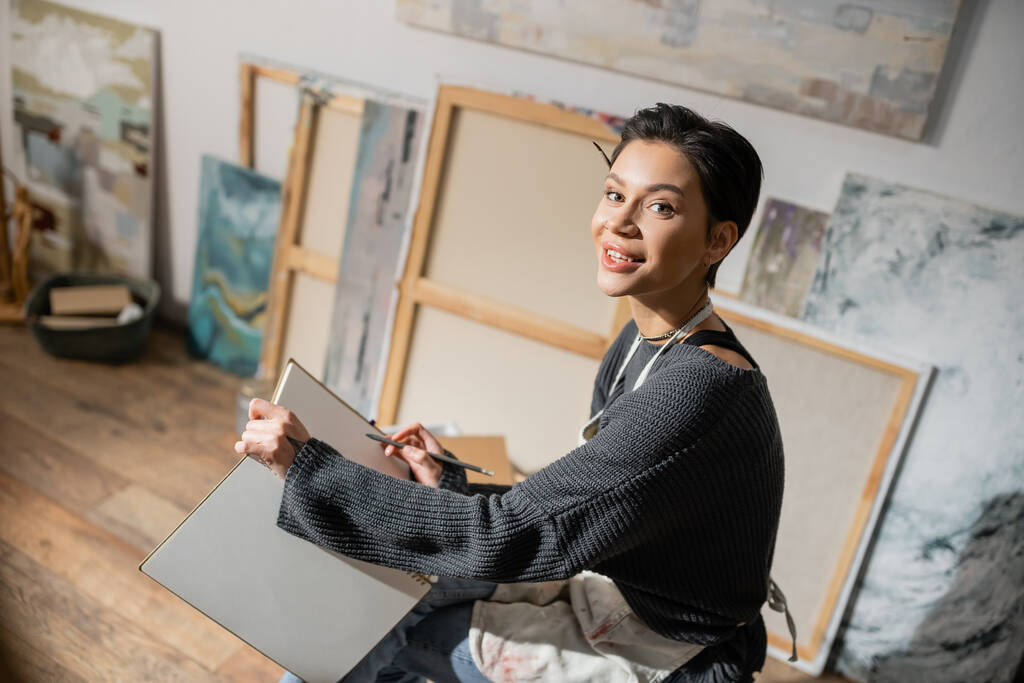 Young short haired artist holding pencil and sketchbook in studio  - Photo, Image