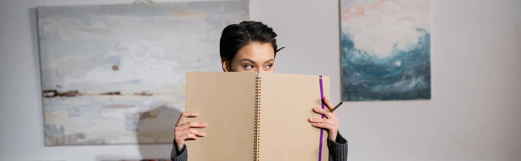 Artist holding pencil and sketchbook near face and looking away in workshop, banner  - Foto, immagini