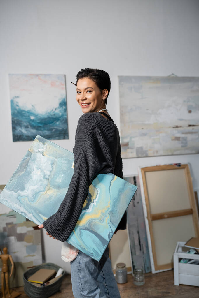 Happy artist looking at camera and holding painting in studio  - Foto, Imagem