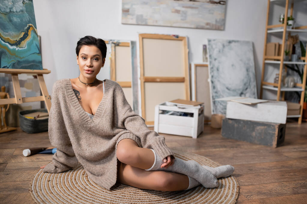 Σέξι καλλιτέχνης σε πουλόβερ κοιτάζοντας κάμερα, ενώ κάθεται στο πάτωμα στο στούντιο  - Φωτογραφία, εικόνα