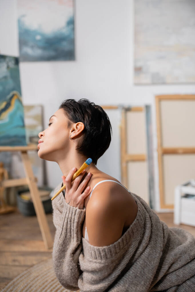 Πλάγια άποψη του κοντά μαλλιά μελαχρινή γυναίκα σε πουλόβερ κρατώντας πινέλο στο στούντιο καλλιτέχνη  - Φωτογραφία, εικόνα