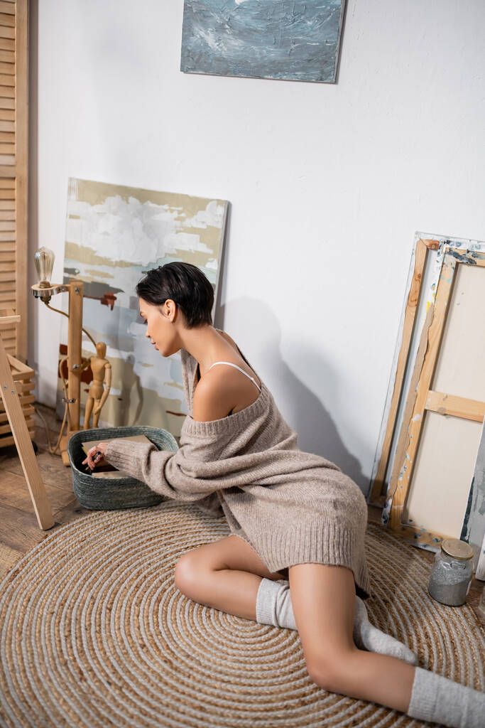 Side view of sexy artist taking paintbrush from basket near paintings in studio  - Fotografie, Obrázek