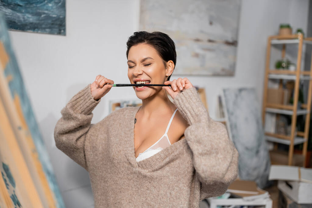 Seductive artist in bra and sweater biting paintbrush in studio  - Photo, Image