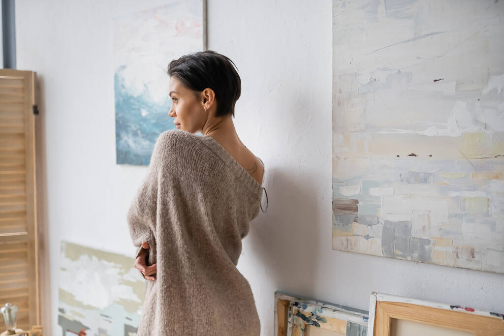 Вид сбоку сексуальной брюнетки в свитере, стоящей рядом с картинами в студии  - Фото, изображение