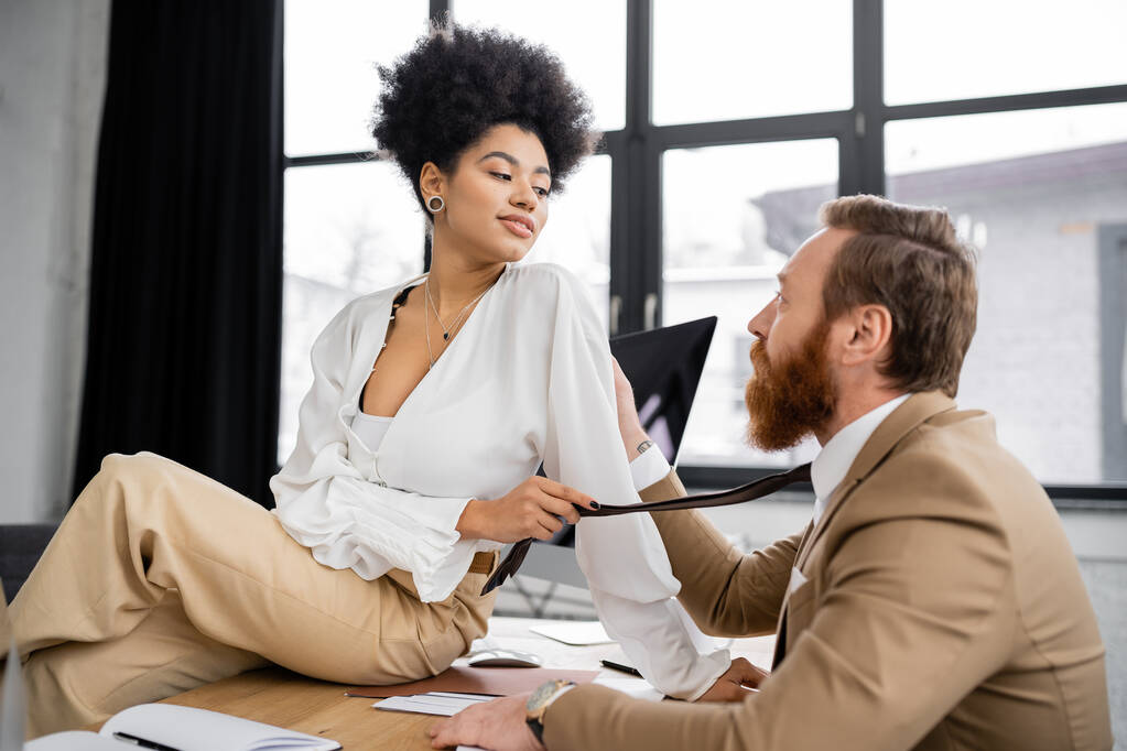 cheerful african american woman sitting on desk and pulling tie of bearded coworker in office  - Foto, Bild