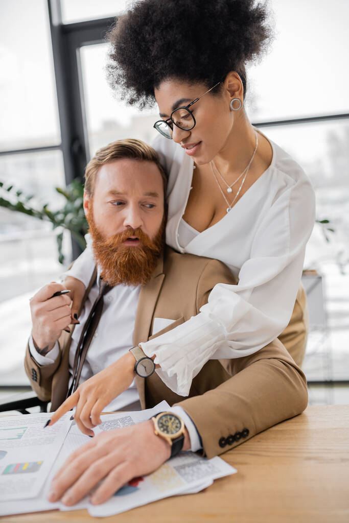 passionate african american woman leaning on shoulders of bearded man while pointing at charts  - Foto, Bild