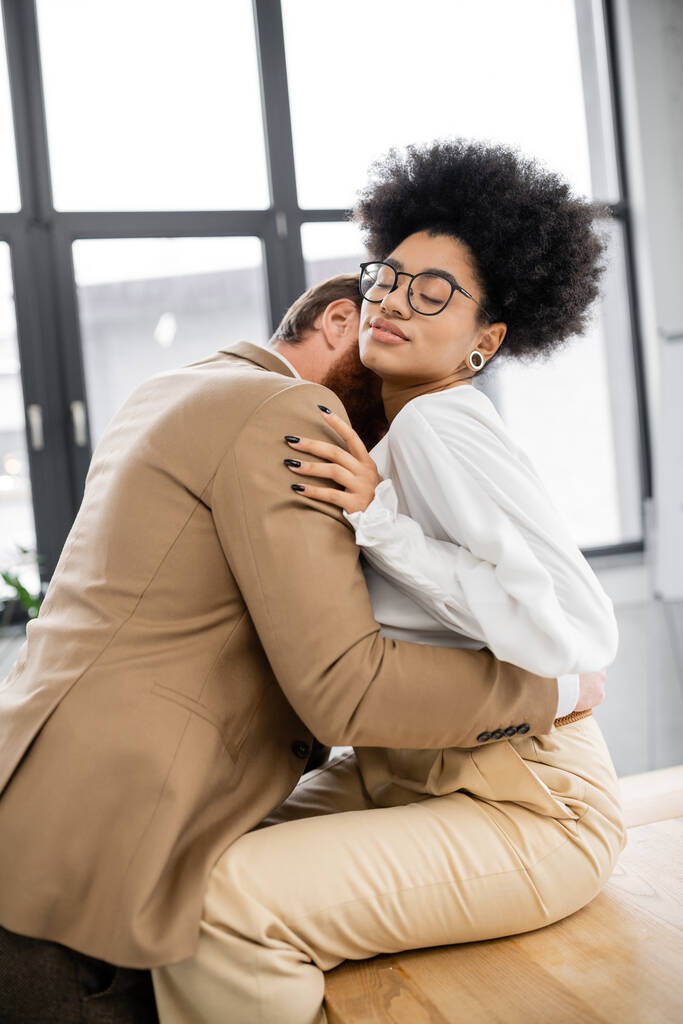 страстный мужчина целует шею кудрявой африканской американки в очках, сидящей на столе в офисе  - Фото, изображение