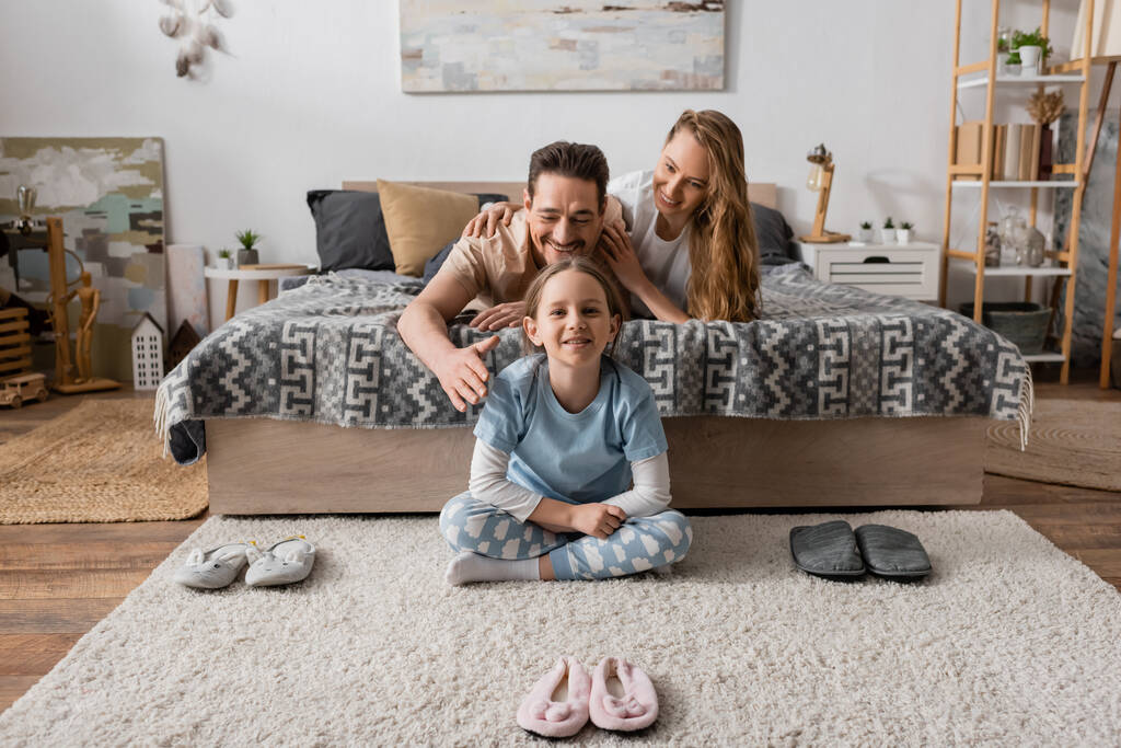 positive child sitting on carpet around slippers near happy parents on bed  - Foto, Bild