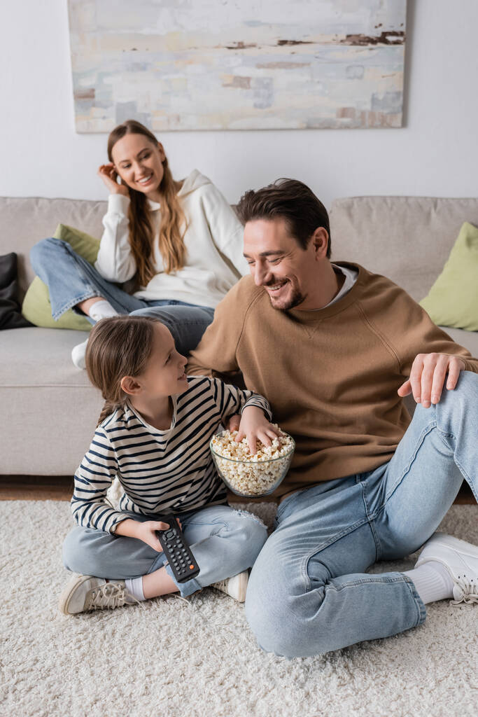 cheerful kid with remote controller looking at father while reaching popcorn near mother on blurred background  - Foto, immagini