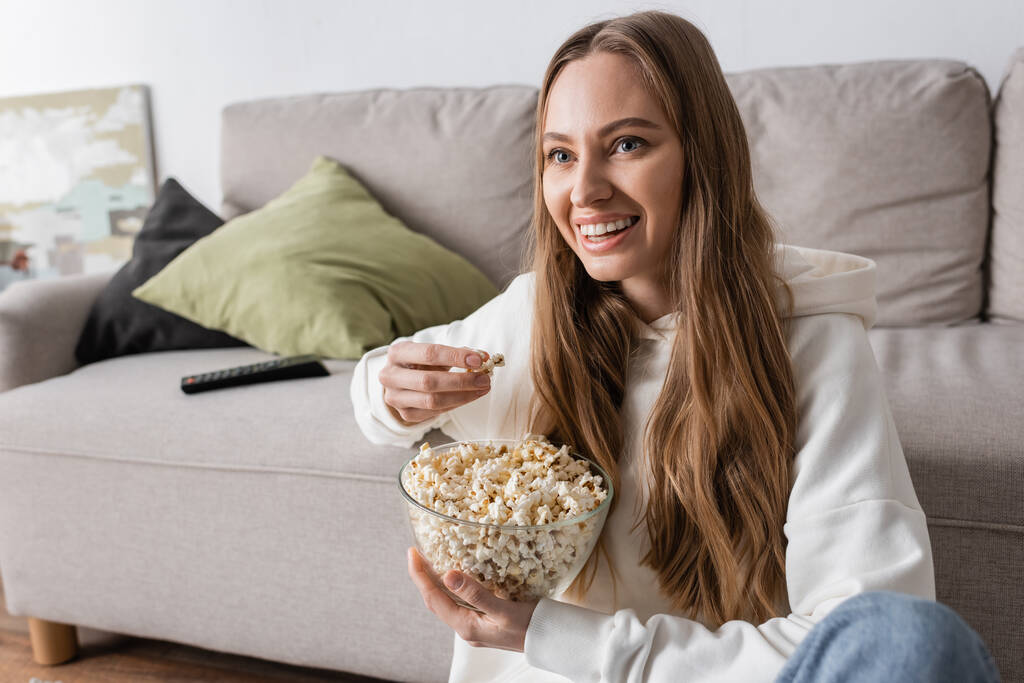 happy woman holding bowl with popcorn while watching movie in living room - Fotó, kép