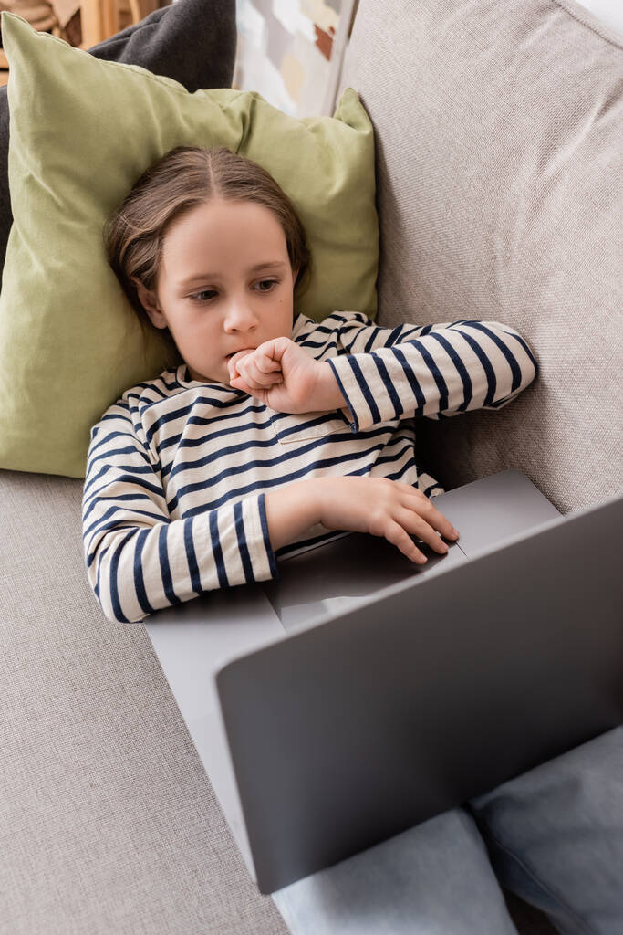high angle view of focused preteen girl in casual clothes using laptop while lying on couch  - Fotografie, Obrázek