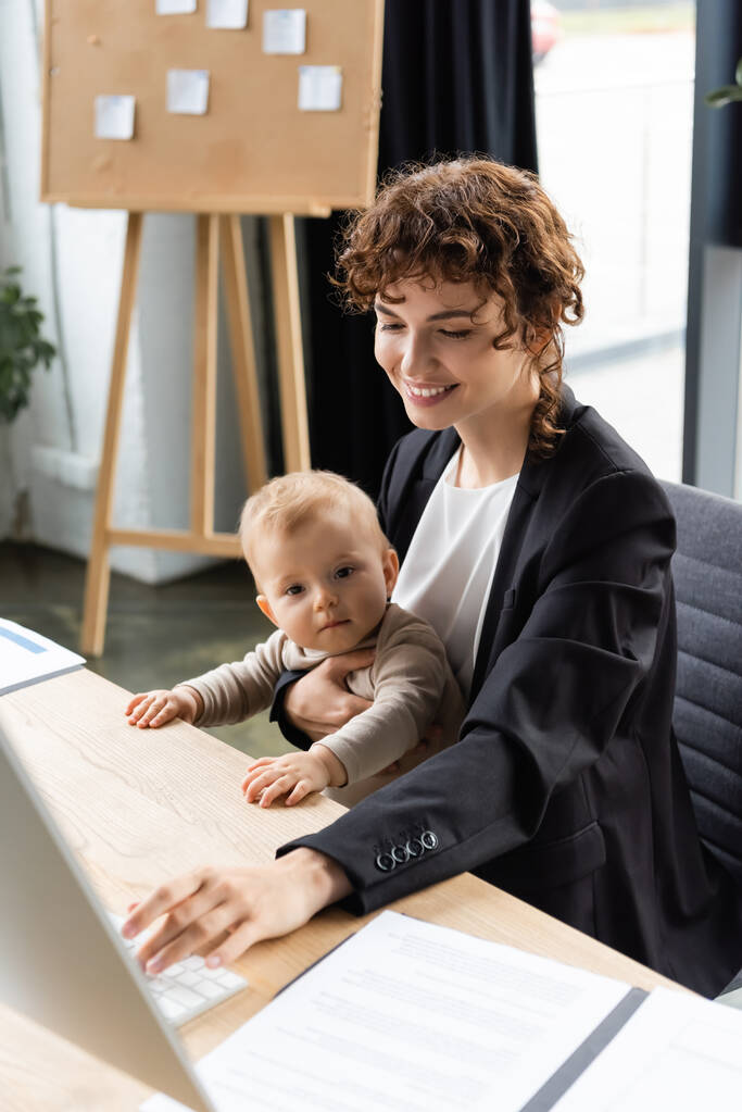 smiling businesswoman working on computer while sitting at workplace with toddler daughter - Foto, afbeelding
